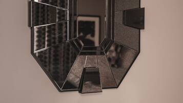 Une œuvre d’art géométrique pour mur, composée d’un miroir central encadré d’un cadre hexagonal. En dessous, une console noire supporte un livre et une paire de ciseaux.