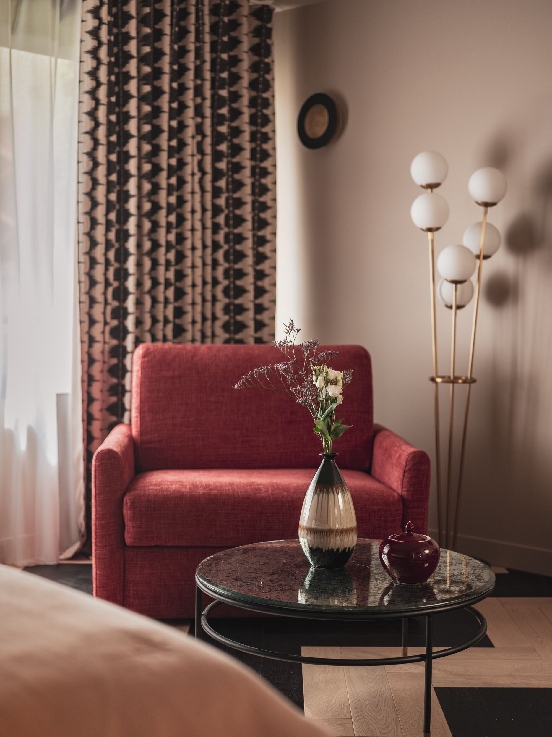 Una habitación con un sillón rojo, una mesa de vidrio con un florero y una lámpara de pie con luces esféricas.