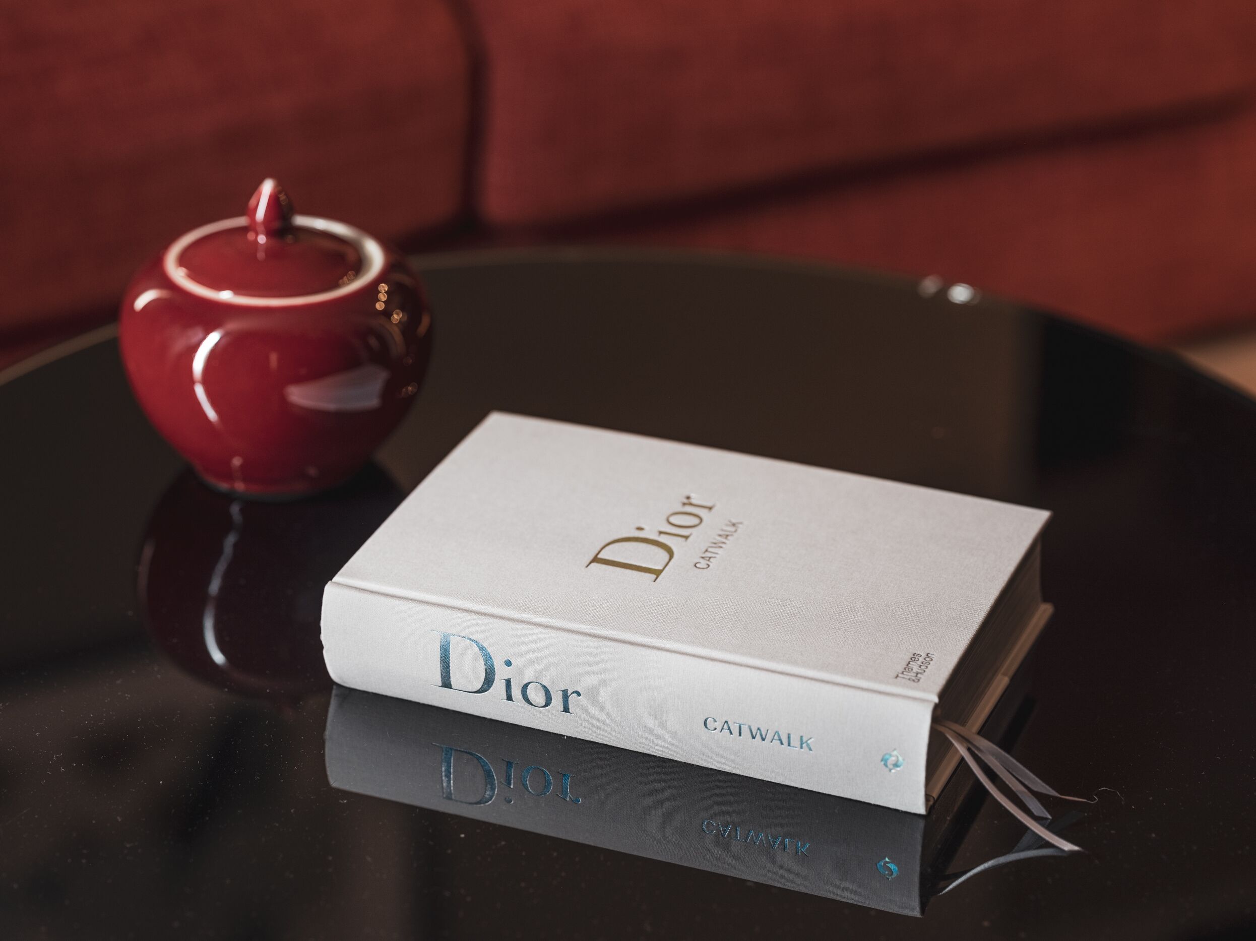 A book titled 'Dior Catwalk' on a black table with a red teapot beside it.