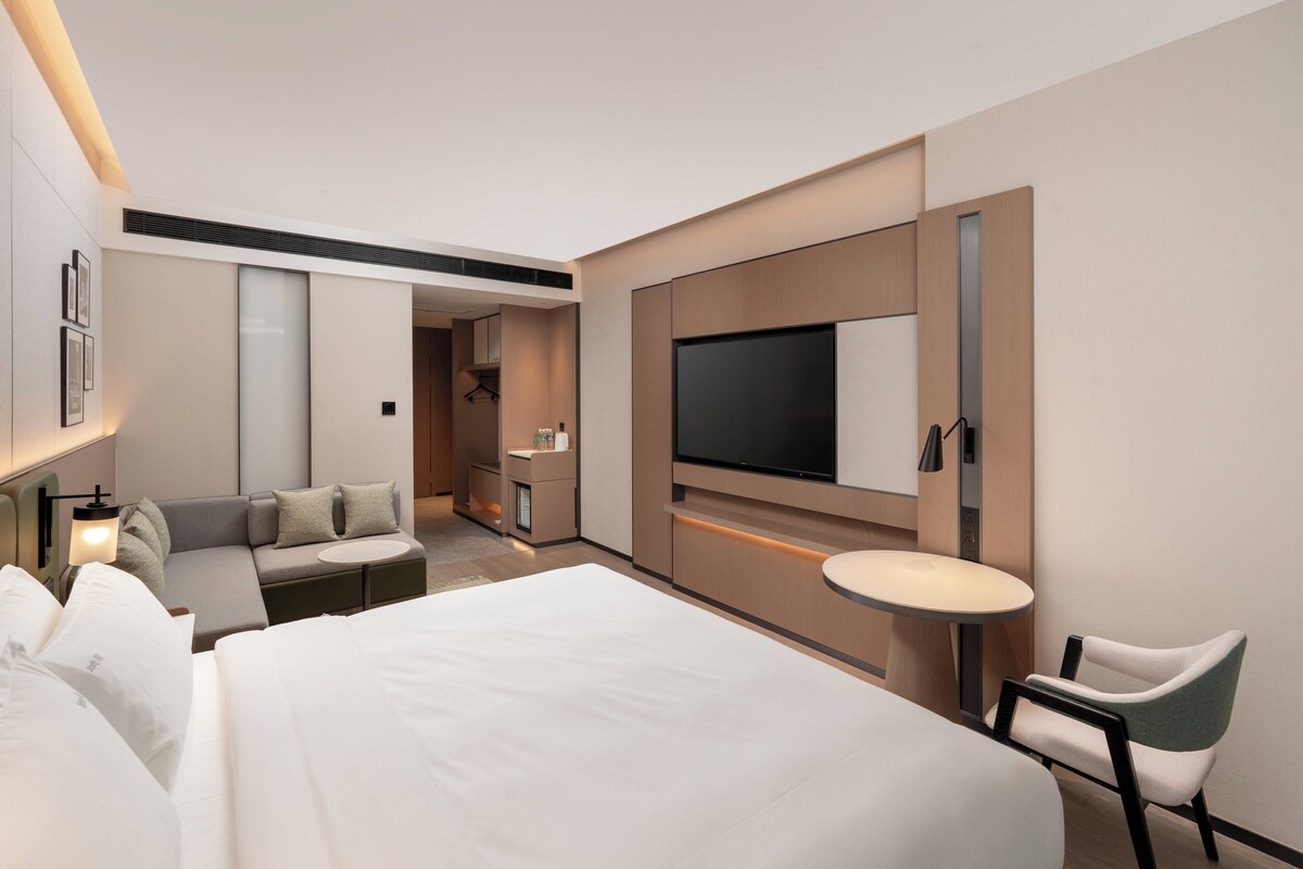 A modern hotel room with a large bed, a sofa, a TV mounted on the wall, a small round table, and a chair.