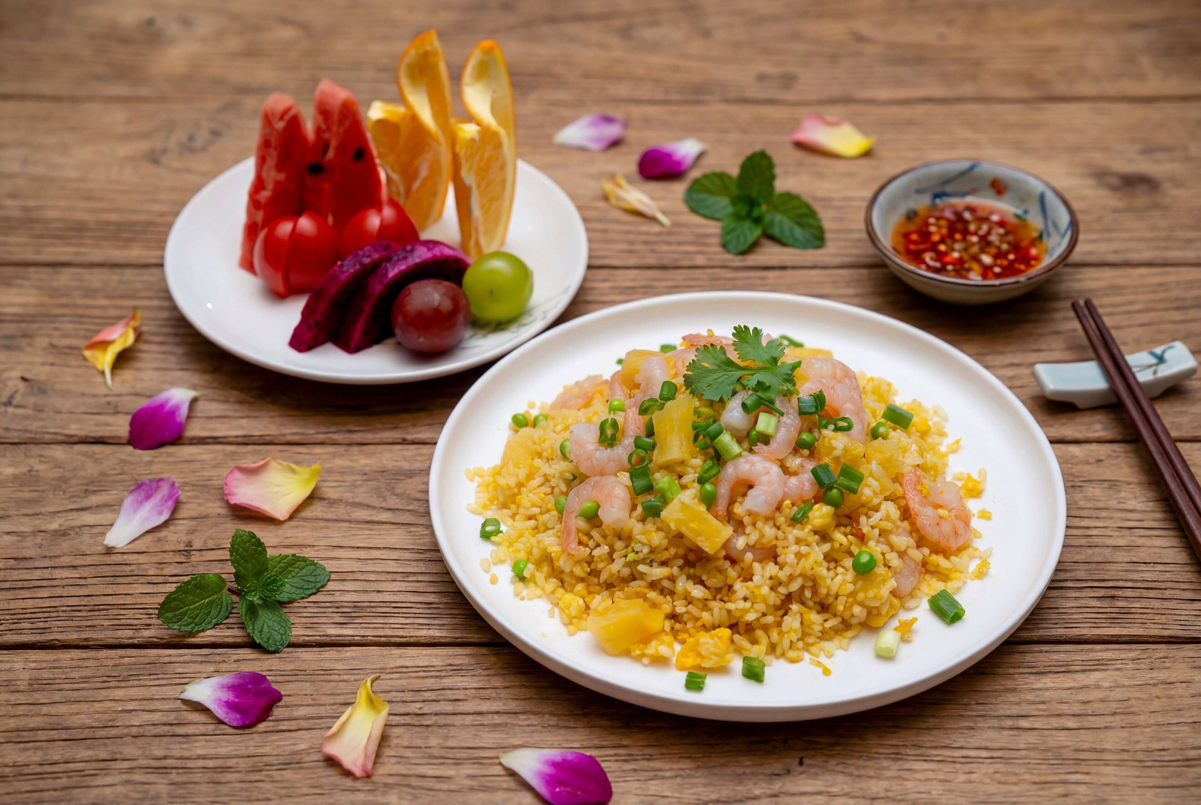 Um prato de arroz frito com camarão, decorado com cebolinha e coentro, acompanhado de melancia e fatias de laranja, e uma pequena tigela de molho de pimenta vermelha.