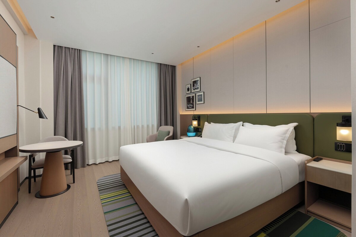 A modern hotel room with a large bed, a desk, and a chair. There is a window with curtains, a nightstand with a lamp, and a wall-mounted TV.