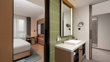Una habitación de hotel moderna con una cama, un escritorio pequeño, un tocador con espejo y un baño con lavabo e inodoro.