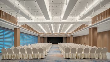A conference room with rows of white chairs, a stage, and a high ceiling with linear lighting fixtures.