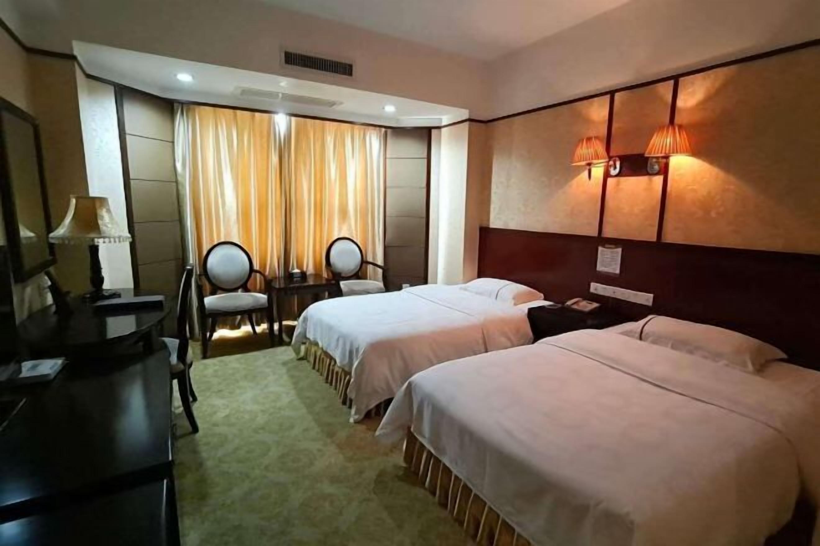 A hotel room with two beds, a desk, a chair, and a lamp.