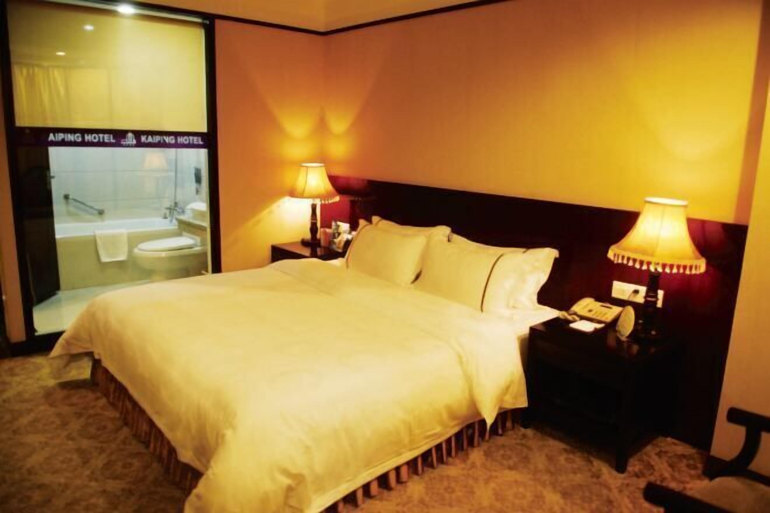 A hotel room with a large bed, two bedside lamps, a nightstand with a telephone, and a view of the bathroom through a glass door.