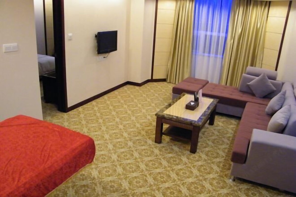 A hotel room with a patterned carpet, a sofa, a small table, a television on the wall, and a bed with a red cover.