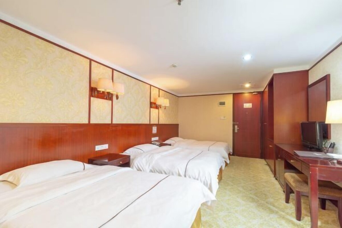 A hotel room with three beds, a desk with a chair, a television, and wall-mounted lamps.