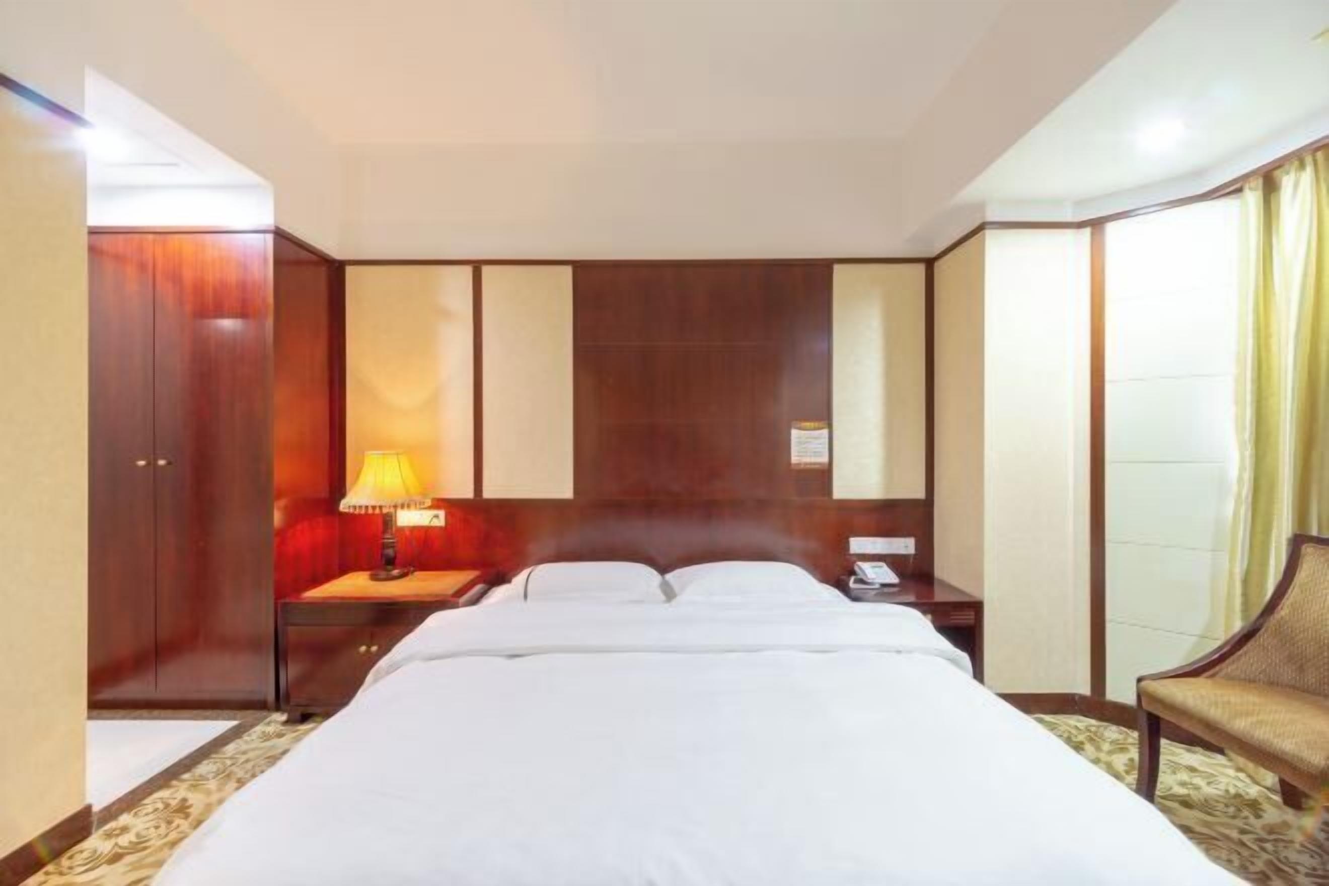 Una habitación de hotel con cama, mesita de noche con lámpara, una silla y un armario.