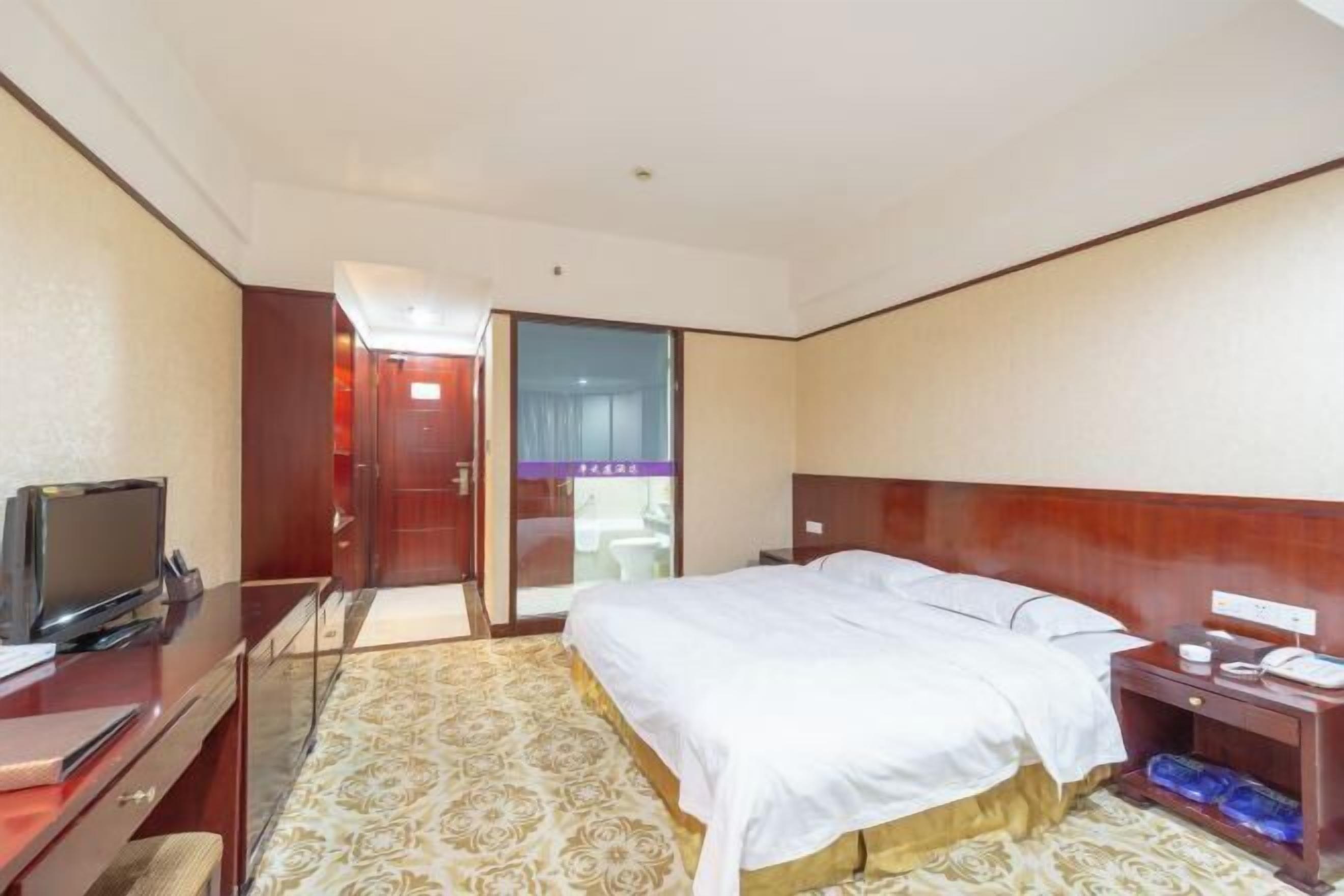 A hotel room with a bed, a desk with a television, a nightstand, and a bathroom visible through a glass door.