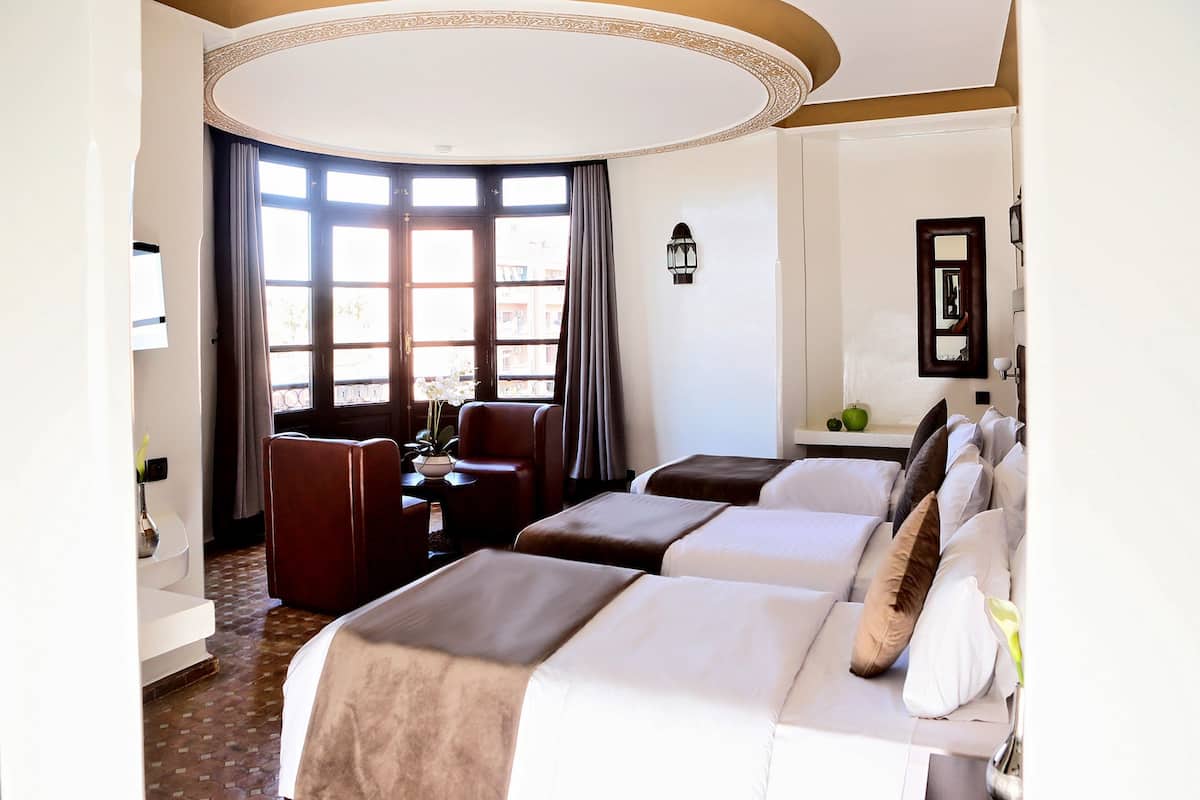 Habitación de hotel con dos camas, un área de descanso con una mesa y sillas, y un ventanal con cortinas.