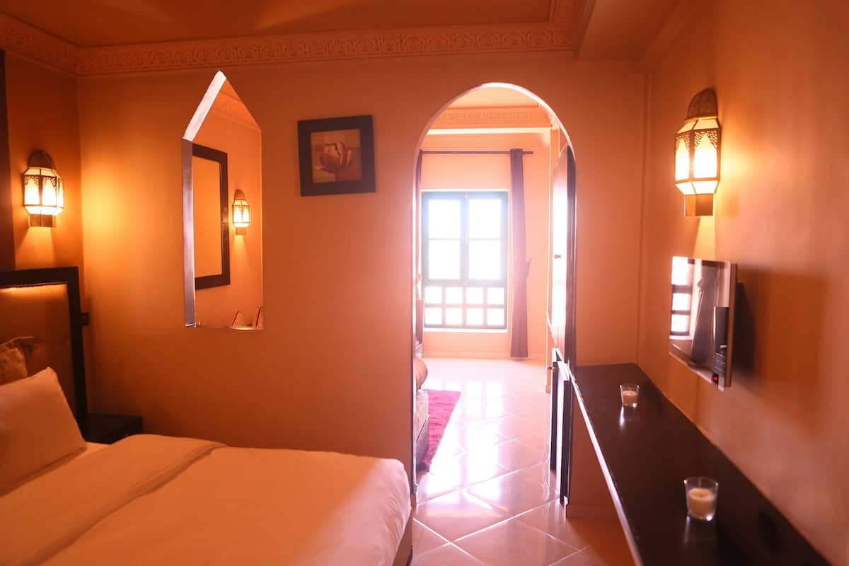 Una habitación de hotel con cama, mesita de noche, espejo, apliques de pared y un umbral que da a otra habitación.
