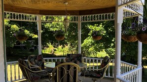 Un espacio al aire libre con techo, equipado con sillas de mimbre y una mesa, rodeado de barandas blancas y macetas colgantes.