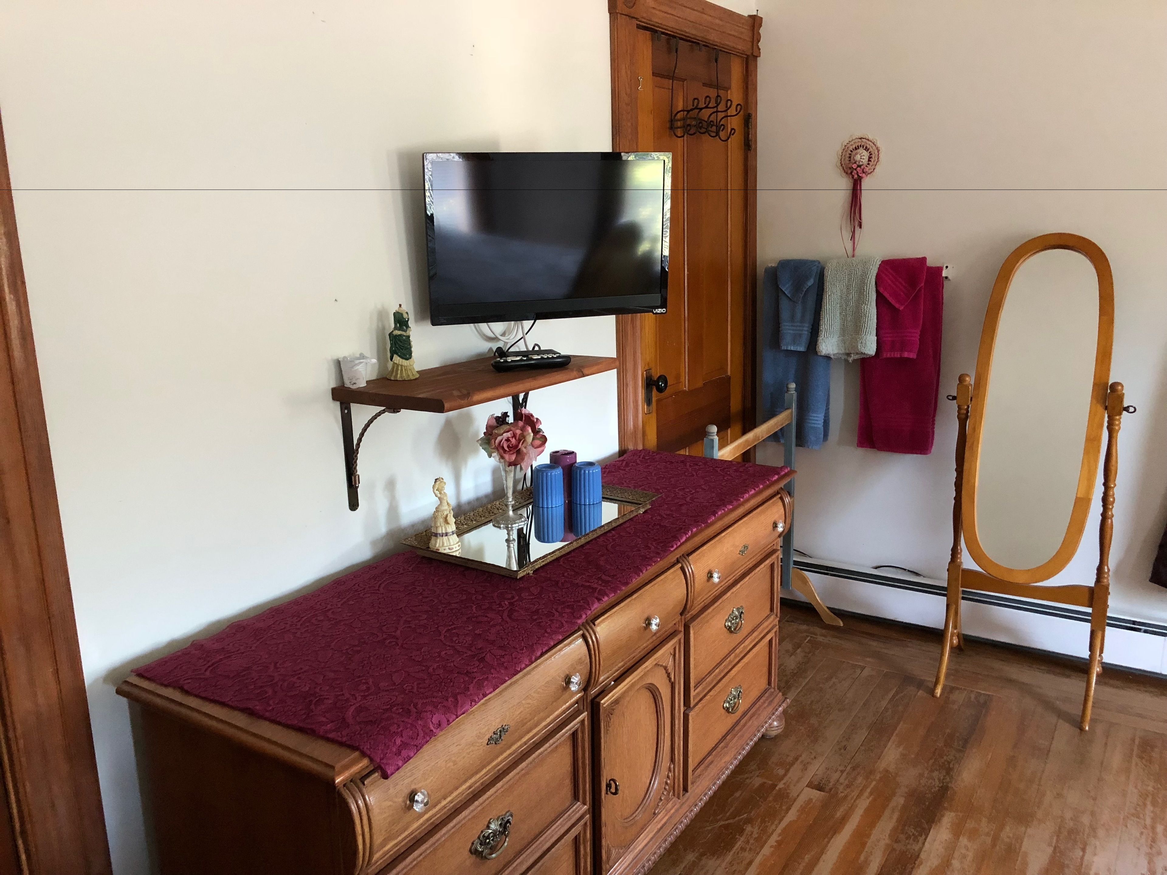 A room with a wooden dresser, a television on a wooden shelf, a vanity with a mirror, and a wooden chair.