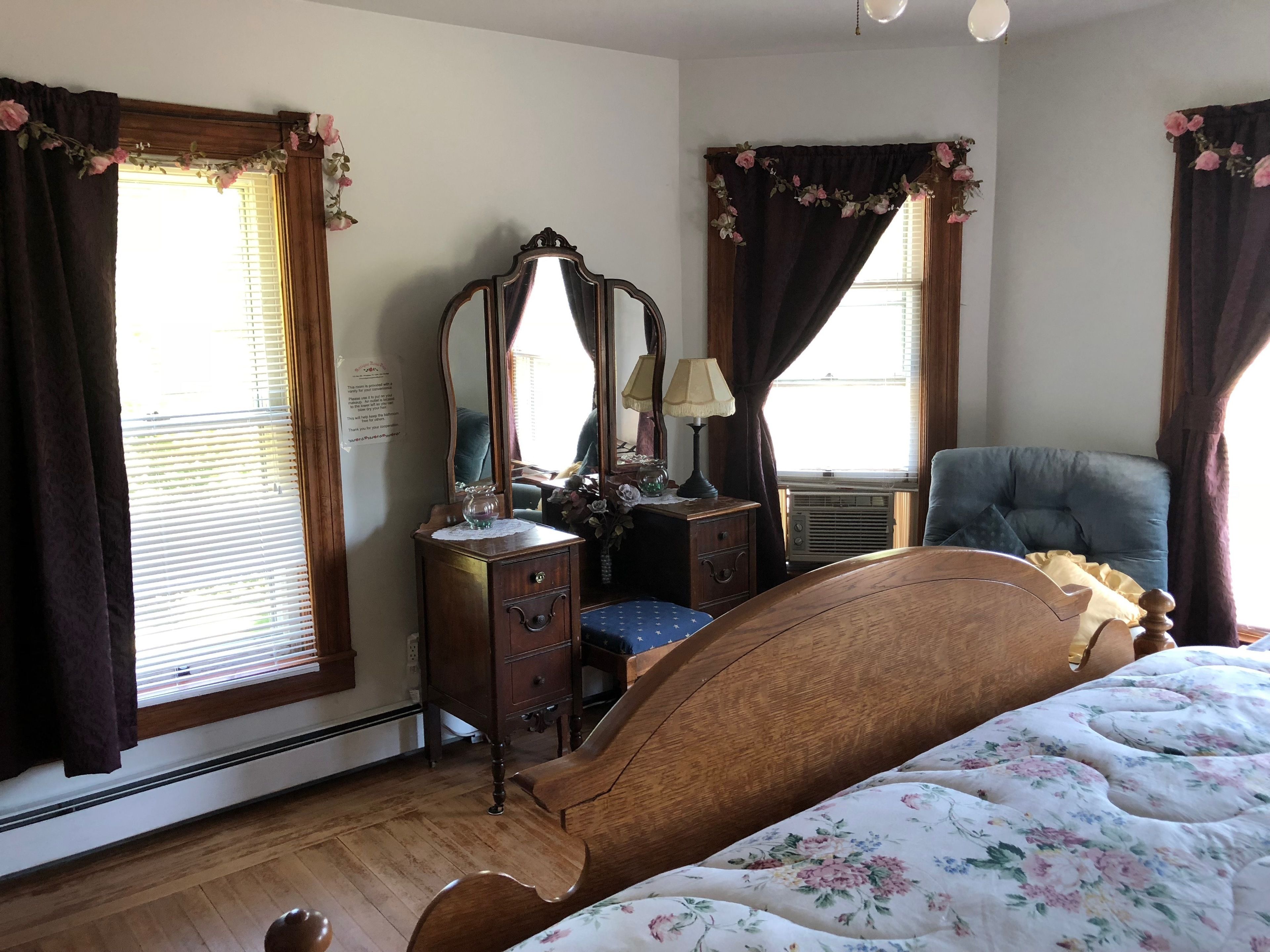 A bedroom with a wooden bed, a dresser with a mirror, a chair, and a window with floral curtains.