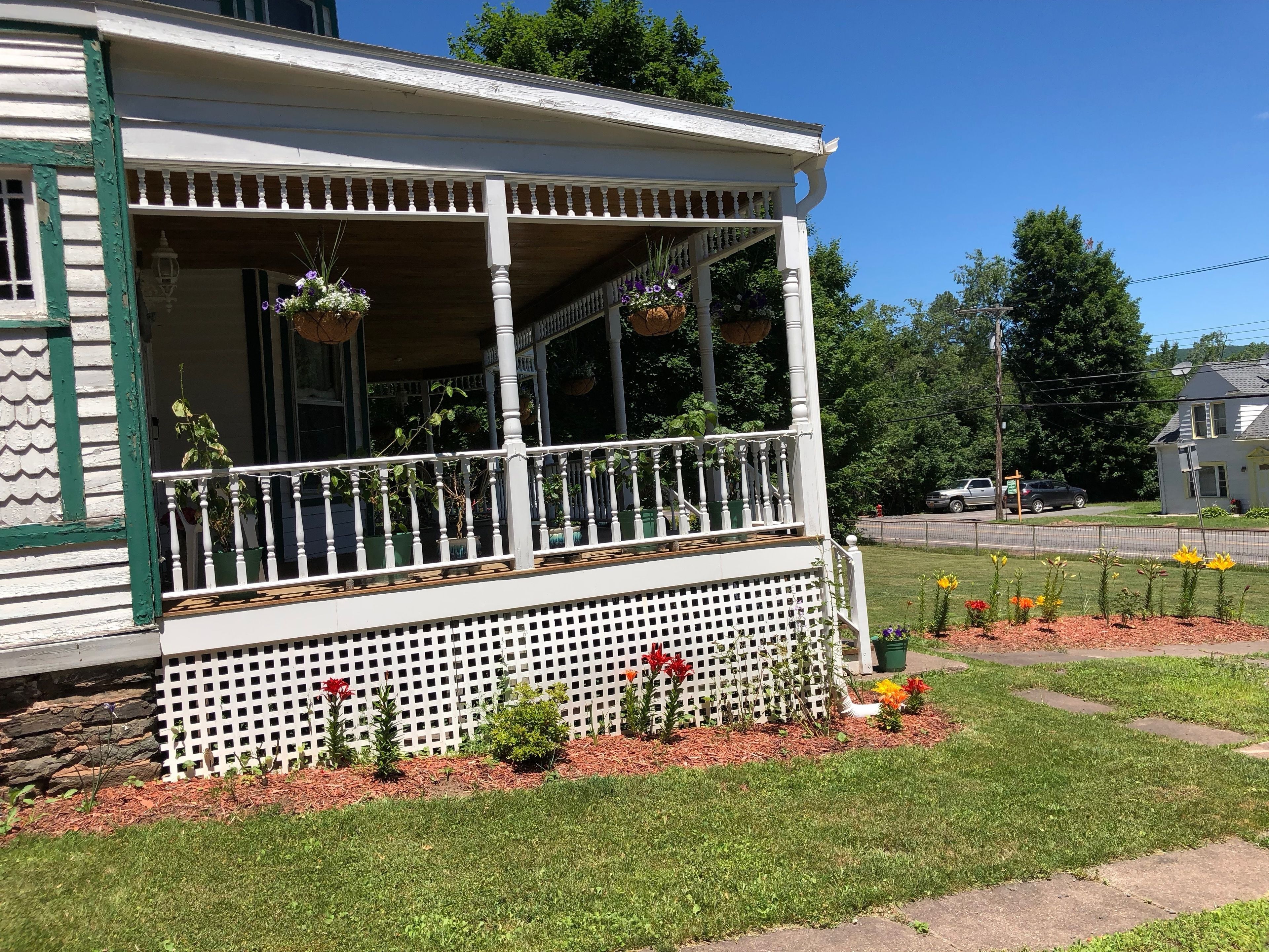 Ein zweigeschossiges Haus mit überdachter Veranda, weißem Zaun und einem Garten mit Blumen und Sträuchern.