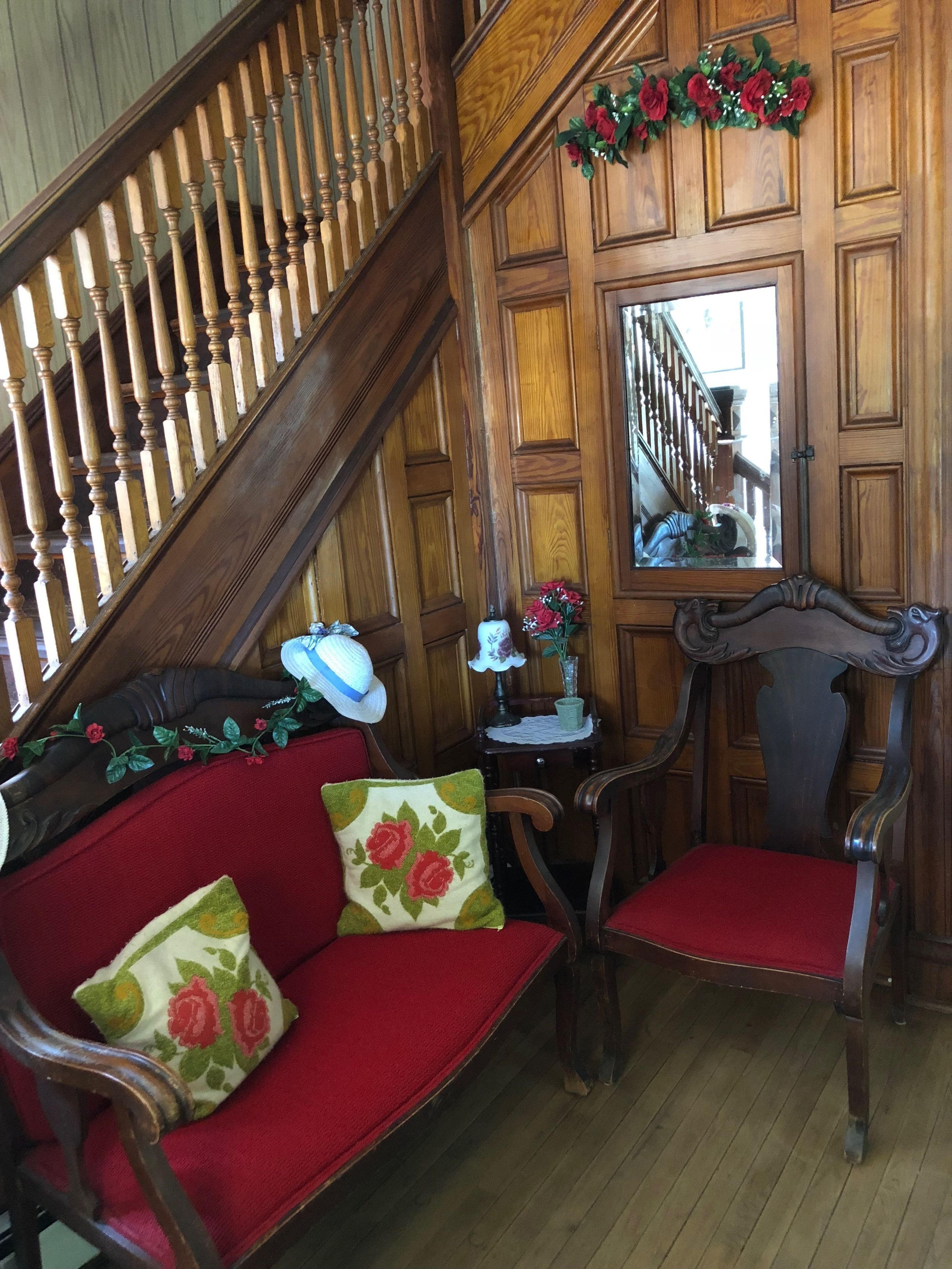 A wooden staircase with a railing, a mirror on the wall, a red sofa with floral cushions, and a wooden chair.