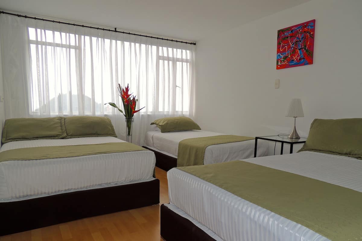 A hotel room with three single beds, a small table with a lamp, and a painting on the wall.