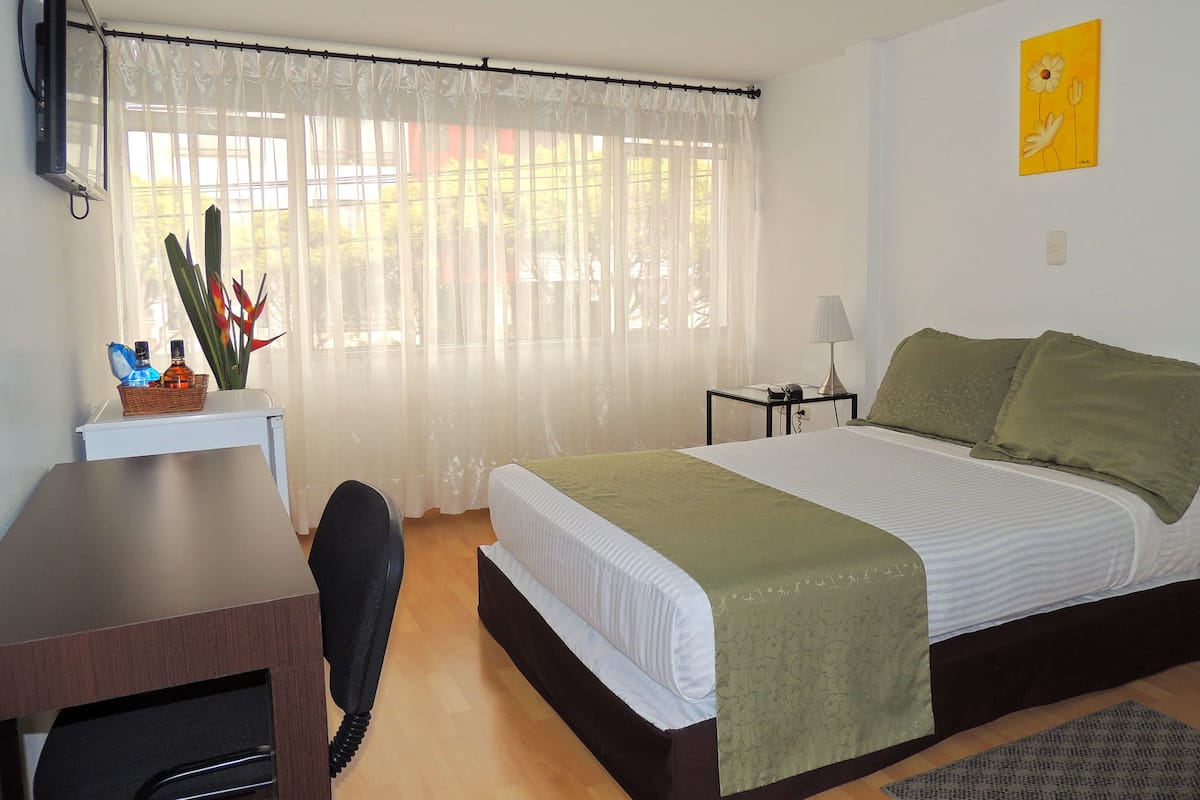 A bedroom with a bed, bedside table, lamp, and a window with sheer curtains.