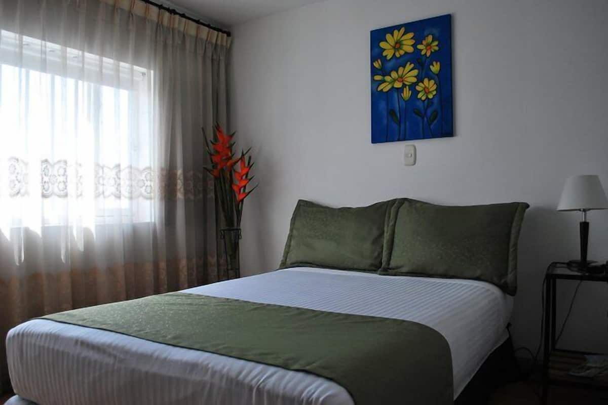 A neatly made bed with a green bedspread, a painting of flowers on the wall, and a vase with red flowers on the windowsill.
