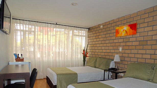 A hotel room with two beds, a desk, and a chair. There is a window with curtains, a brick wall, and a painting. - Hotel Portal del Milán (Manizales)