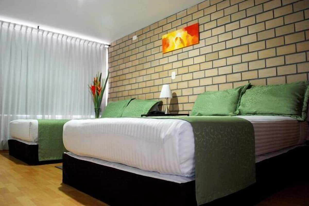 A hotel room with a brick-patterned wall, a bed with green bedding, a bedside table with a lamp, and a window with white curtains.