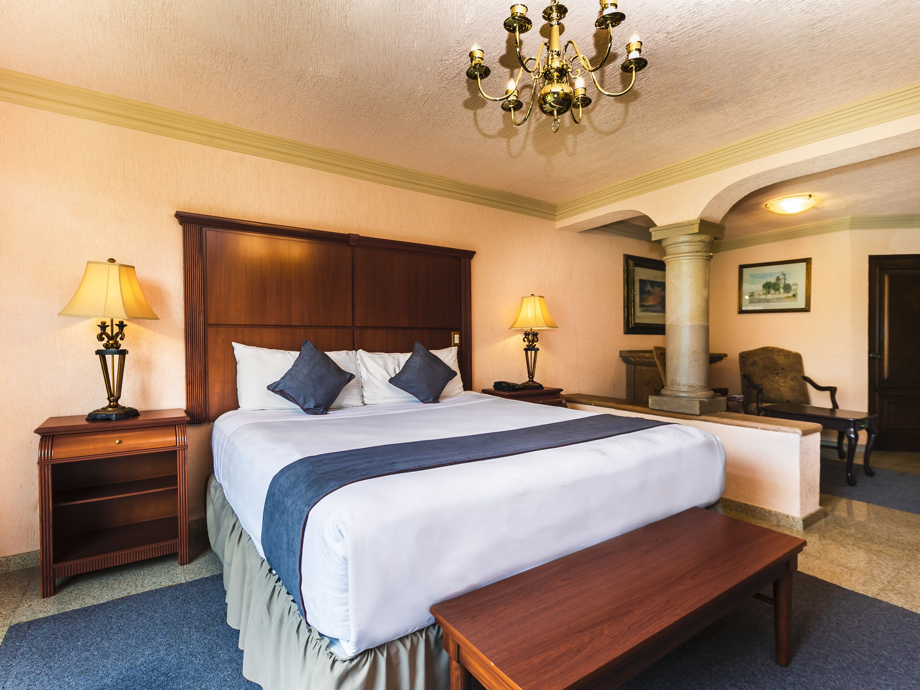 A hotel room with a large bed, two bedside tables with lamps, a bench, a chair, and a chandelier.