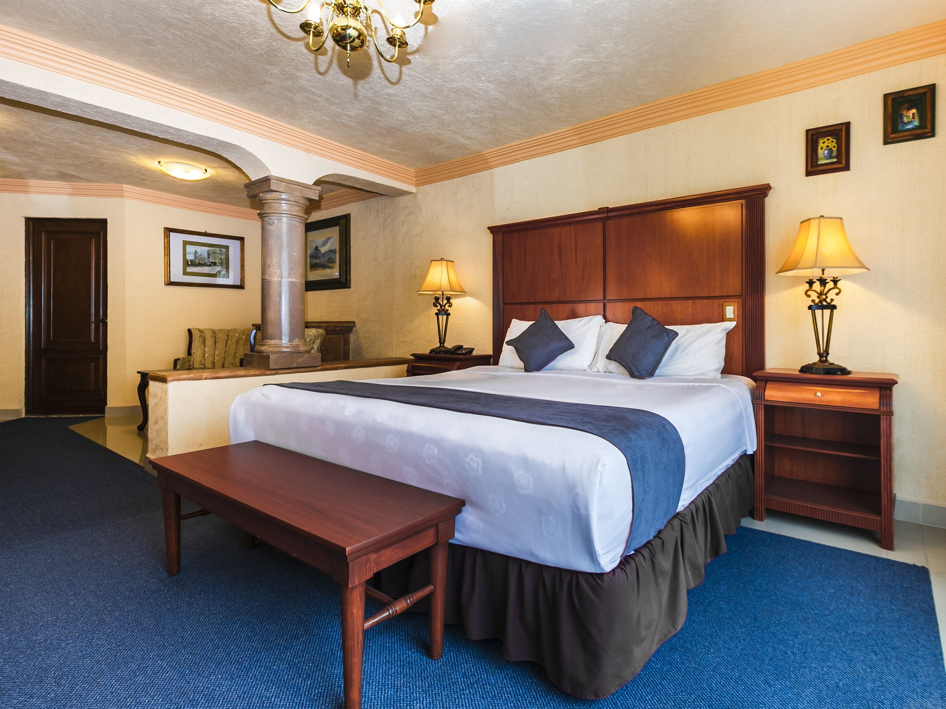 A hotel room with a large bed, two bedside tables with lamps, a wooden bench, a large pillar, and a framed picture on the wall.