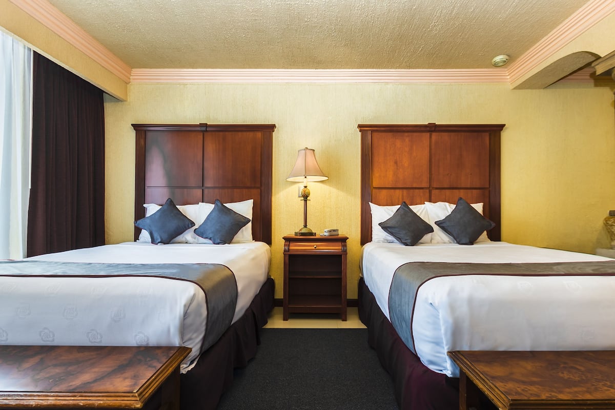 A hotel room with two beds, a nightstand, a lamp, and a window with curtains.