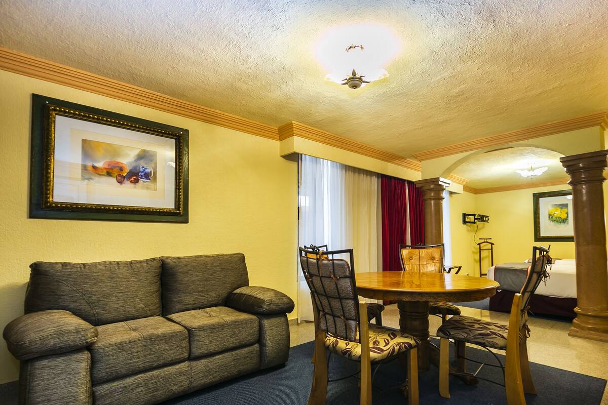 A hotel room with a dining table, chairs, a sofa, a ceiling fan, and a framed picture on the wall.
