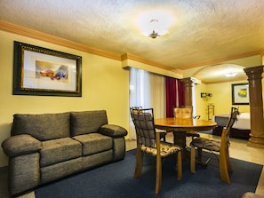 A hotel room with a dining table, chairs, a sofa, a ceiling fan, and a framed picture on the wall.