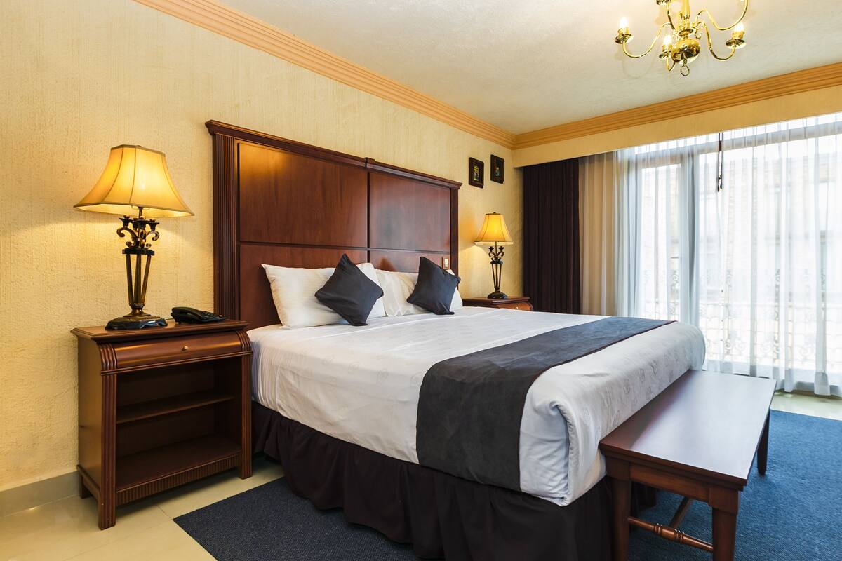 A hotel room with a large bed, two bedside tables with lamps, a wooden headboard, a bench, and a large window with curtains.
