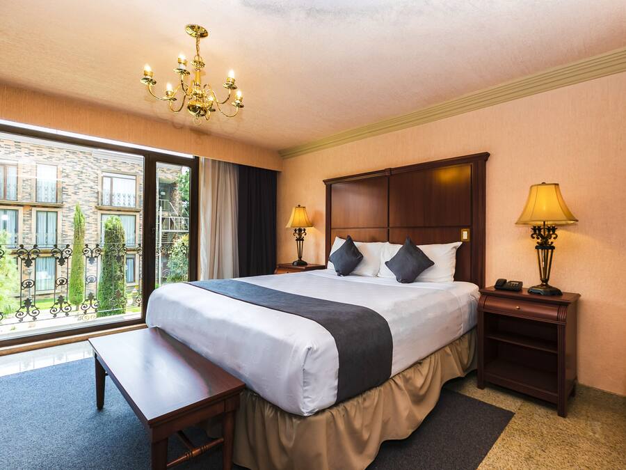 Habitación de hotel con una cama grande, dos lámparas de noche, una mesita de noche de madera, un banco y vistas a un edificio y zonas verdes por la ventana.