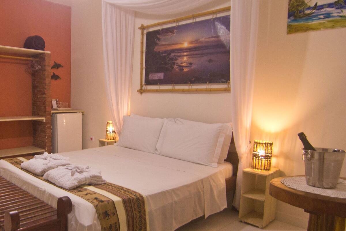 A hotel room with a bed, a bench, a bathroom with a mirror, and a painting on the wall.