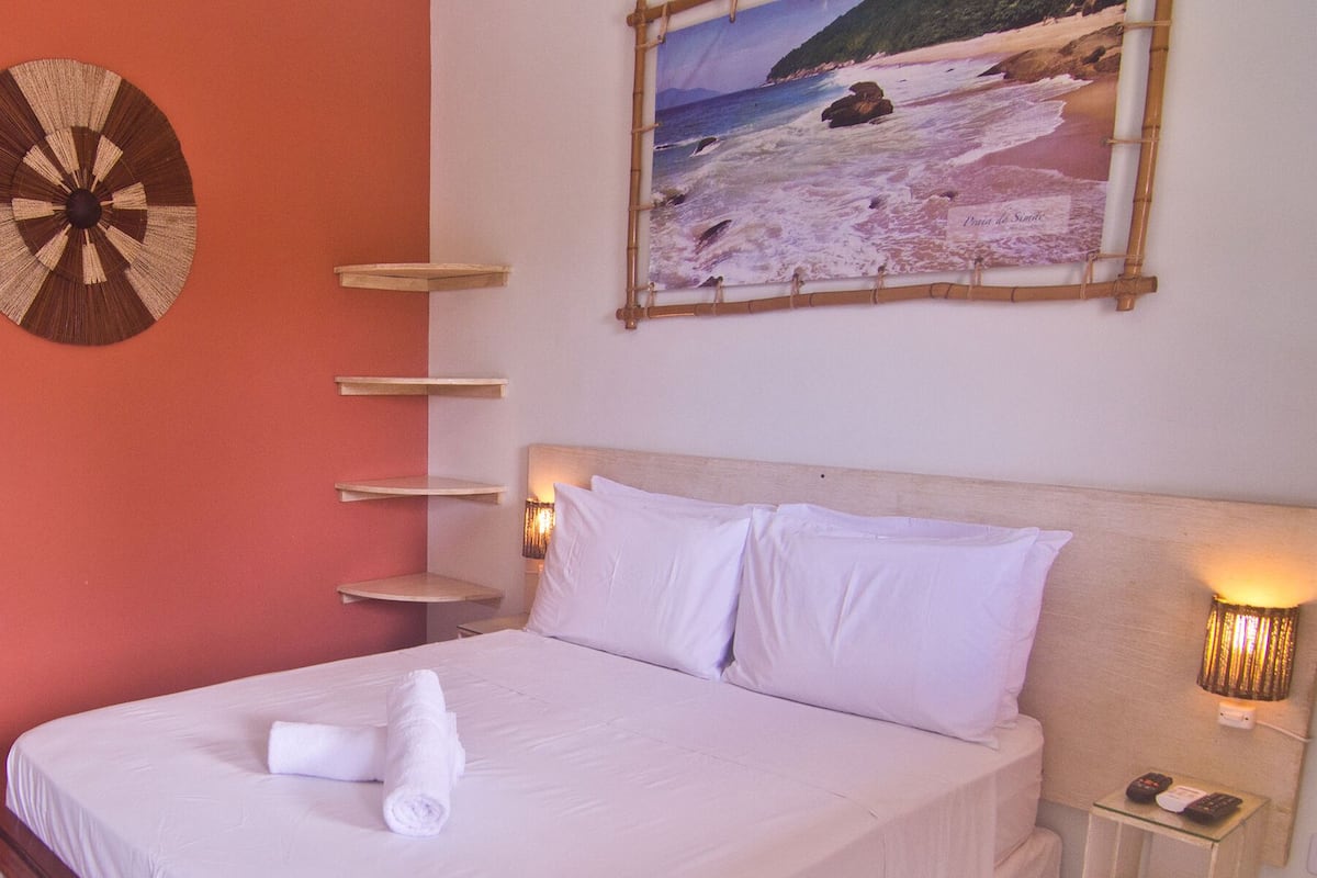 A hotel room with a bed, a wall clock, a painting of a beach, and a small refrigerator.