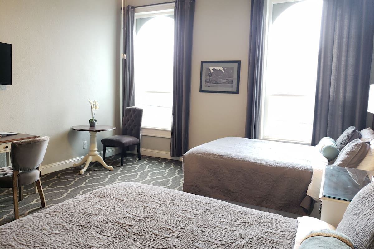 A hotel room with two beds, a desk, a chair, and a small table with a plant.