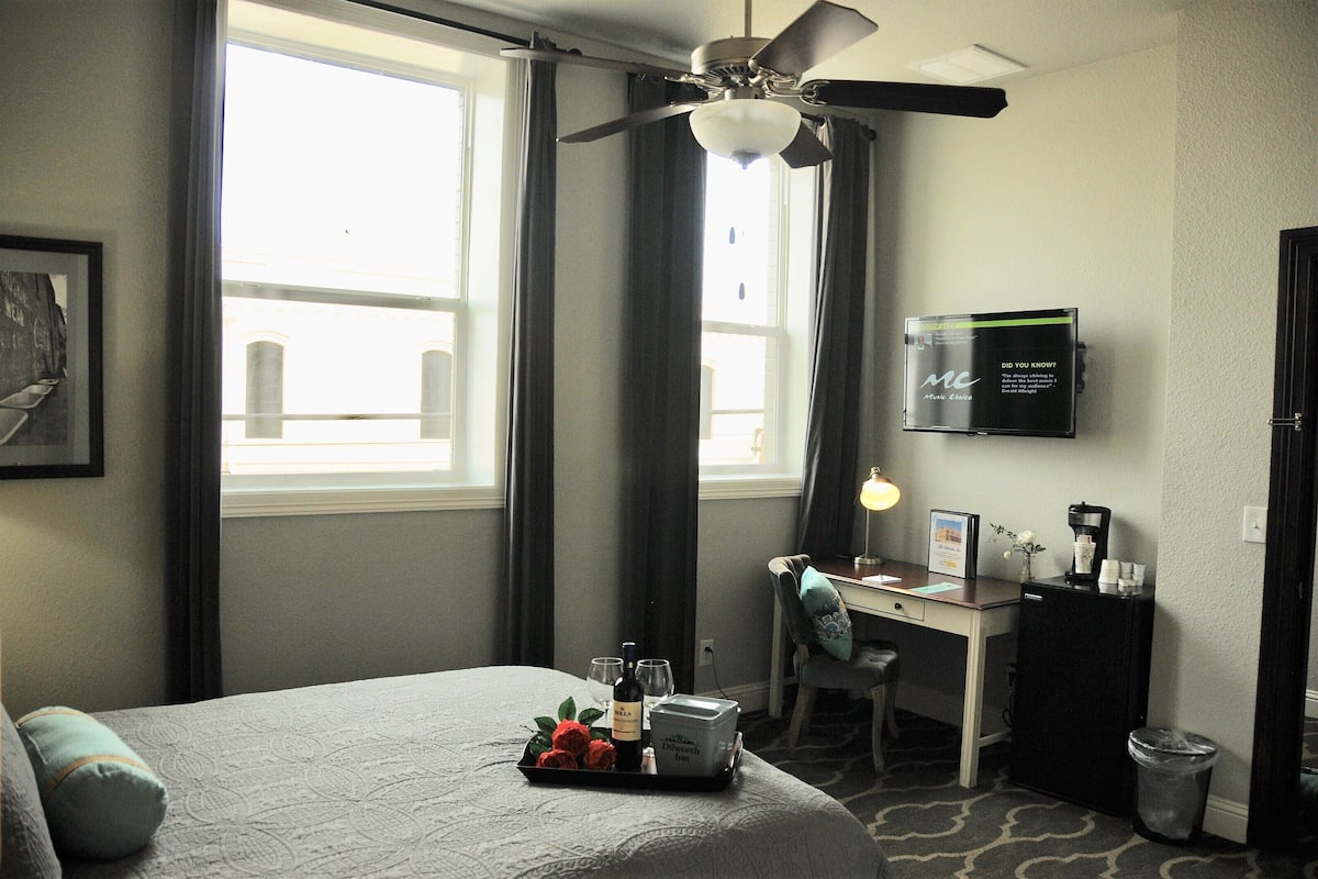 A hotel room with a bed, a desk, a ceiling fan, a TV, and a window with curtains.