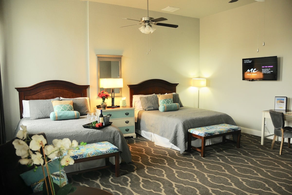 A hotel room with two beds, a desk, a chair, a TV, and a ceiling fan.