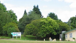 A motel sign with a logo, surrounded by lush greenery and a building with a green roof. - Glowworm Motel (Hangatiki)