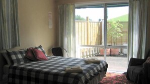 A hotel room with a checkered bedspread, a chair, and a view of a fenced garden through a sliding glass door. - Glowworm Motel (Hangatiki)