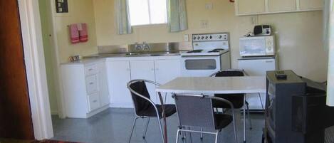 Cocina pequeña con armarios blancos, estufa, microondas y televisor sobre un mueble bajo. Hay dos sillas alrededor de una mesa pequeña.