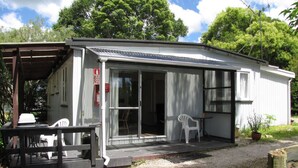 A small, single-story building with a covered porch, a sliding glass door, and a small patio area with a table and chairs. - Glowworm Motel (Hangatiki)