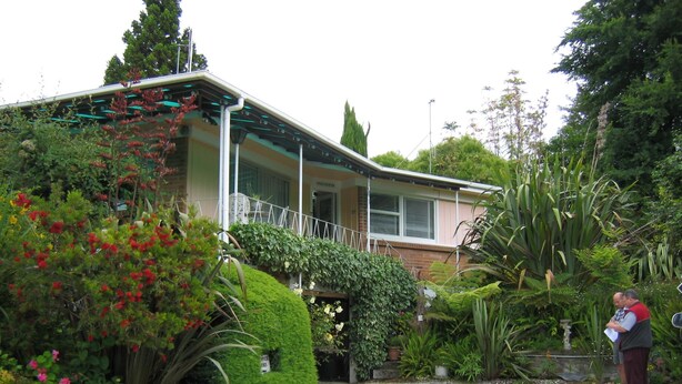 A two-story house with a covered porch, surrounded by lush greenery and a gravel driveway. - Glowworm Motel (Hangatiki)