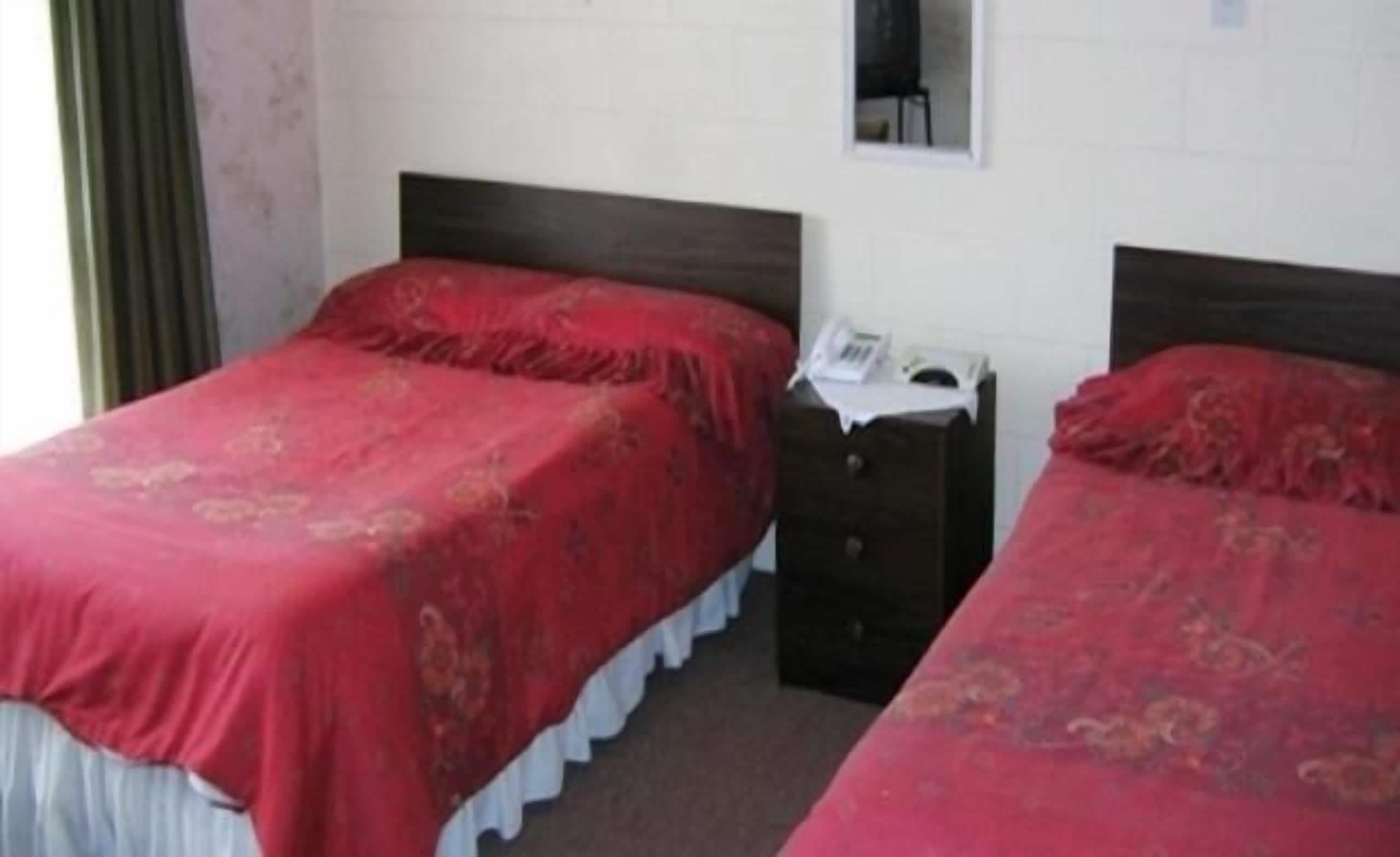 Una habitación con dos camas individuales, cada una con una colcha roja y un cojín blanco. Entre las camas hay una mesita de noche de madera con una lámpara y un teléfono. Las paredes son blancas, y hay una ventana con cortina en el lado izquierdo.