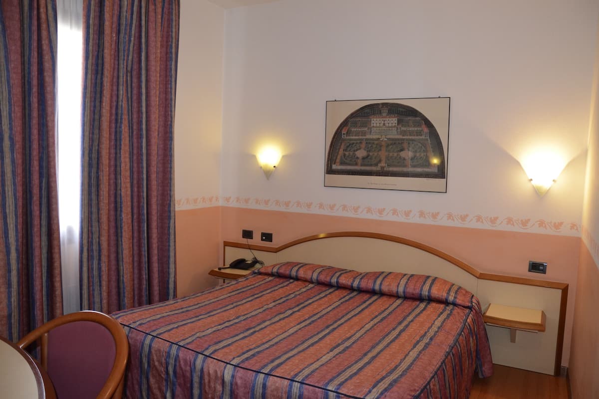 A hotel room with a bed, a nightstand, a chair, and a wall-mounted lamp.