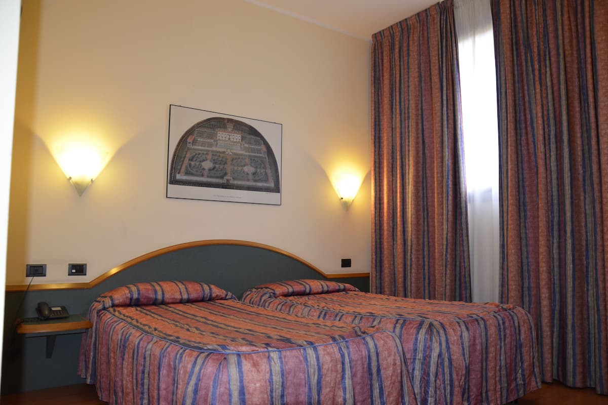 A hotel room with a bed, two wall-mounted lamps, a framed picture, and a window with curtains.