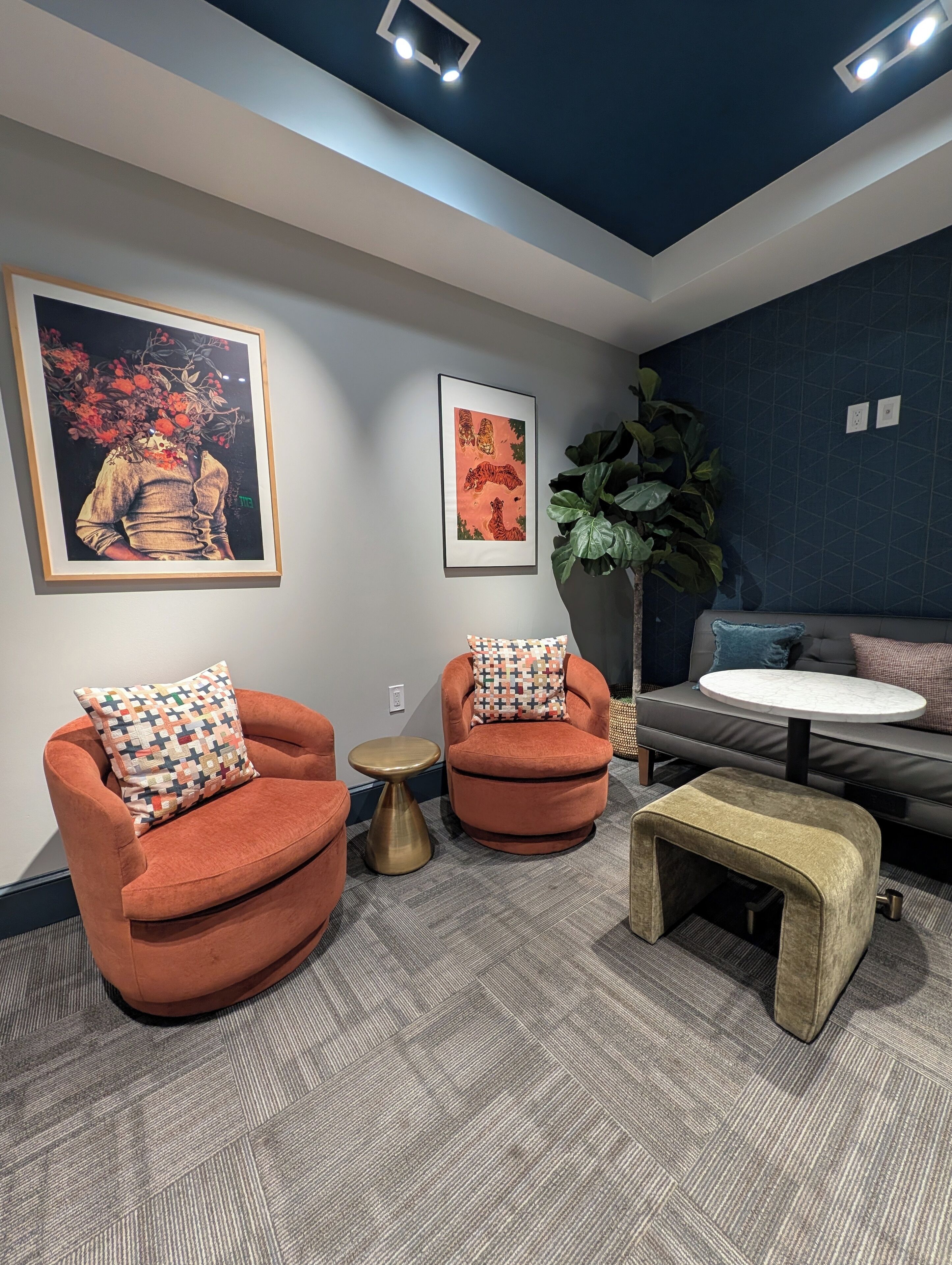 A modern lounge area with two orange armchairs, a round coffee table, and a sofa. There are two framed artworks on the walls and a large potted plant.