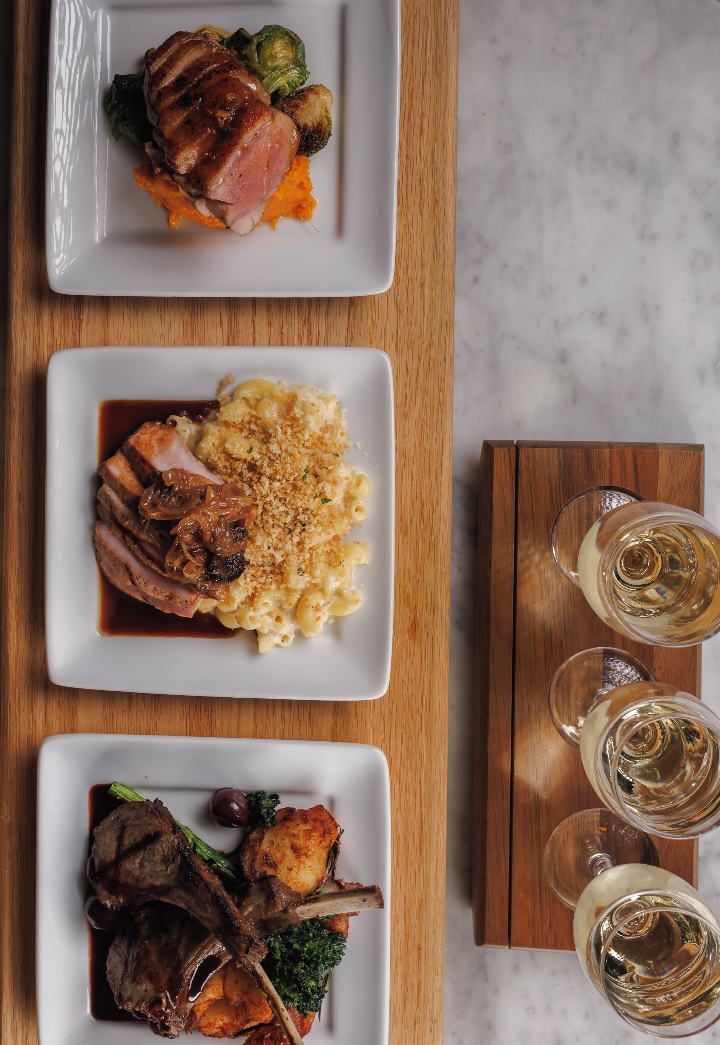 Três pratos de comida sobre uma mesa de madeira, acompanhados de dois copos de vinho branco.