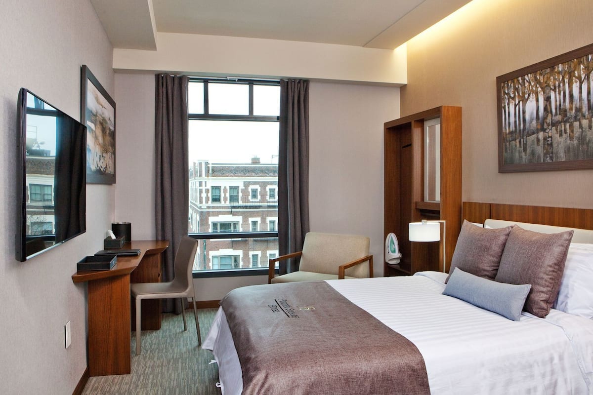 Un cuarto de hotel con cama, escritorio, silla, ventana con vista a edificios y un cuadro en la pared.