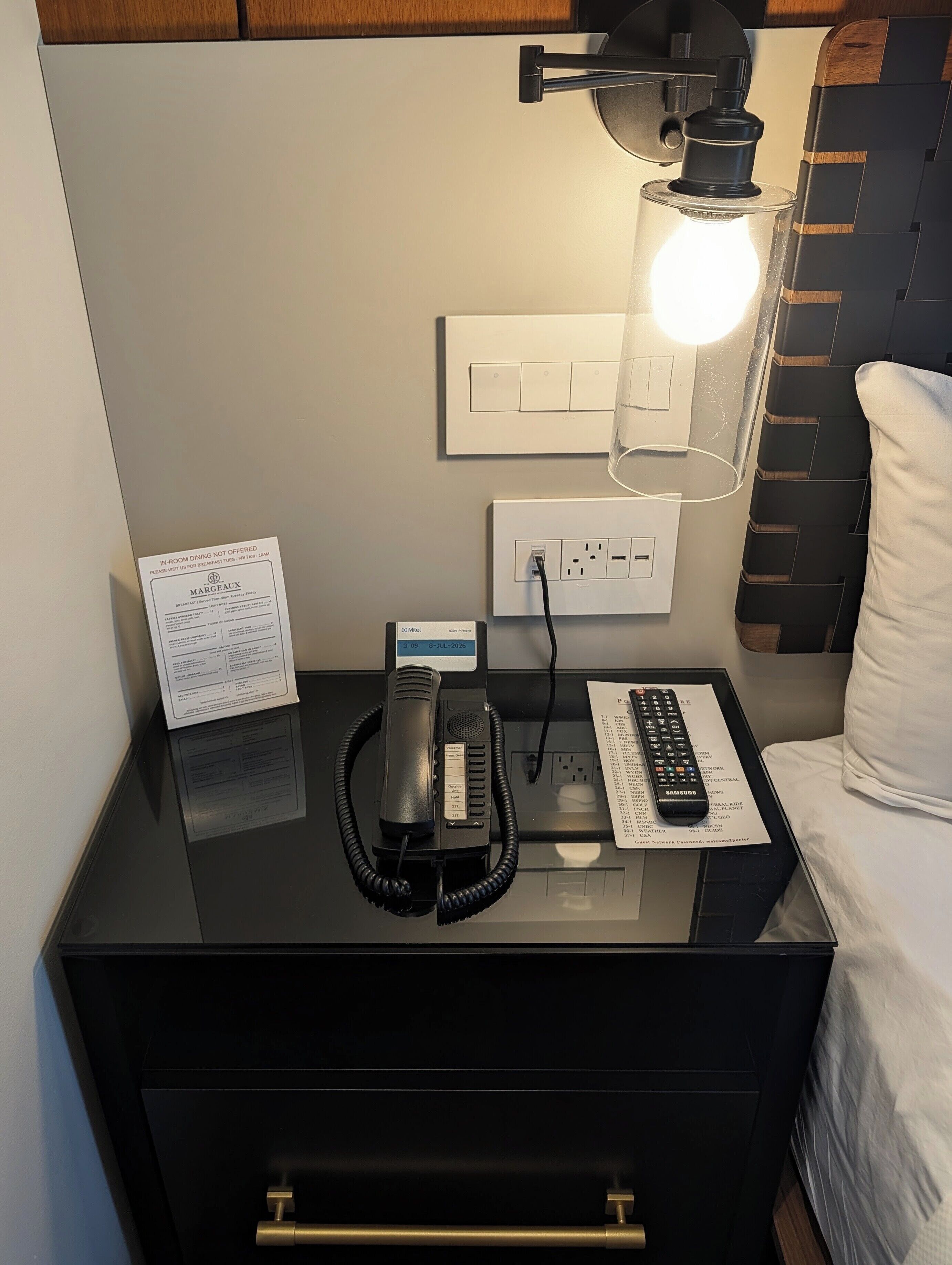 A hotel room with a black bedside table, a lamp, a telephone, a remote control, and a bed with white bedding.